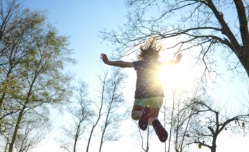 Photo of a little girl jumping high (because trampoline), trees nearby and a sunny day